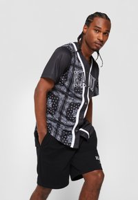 Sort jersey skjorte med hvide knapper og kanter, med et bandana mønster. Kombineret med sorte shorts, der viser en afslappet pasform.