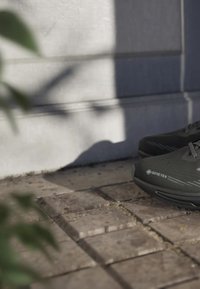 Nærbillede af sort Gore-Tex-sko på udendørs stenbelægning med sollys og skygger på en betonvæg i baggrunden.