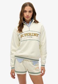 Crèmekleurige sweatshirt met kwartrits heeft het "SUPERDRY" logo en groene accenten, gedragen over een blauw gestreept shirt; gecombineerd met crèmekleurige shorts.