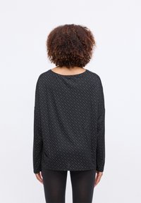 Pessoa com cabelo encaracolado, vestindo uma blusa de manga longa com padrão escuro e leggings pretas, de pé, de costas, contra um fundo branco.