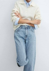 Persona con los brazos cruzados, vistiendo un suéter de punto crema sobre una camisa de cuello azul claro y jeans de mezclilla azul claro de corte holgado, sobre un fondo blanco.