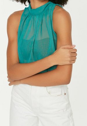 Femme portant un chemisier transparent sans manches bleu sarcelle à col haut et un pantalon blanc, bras croisés sur le torse devant un fond uni.