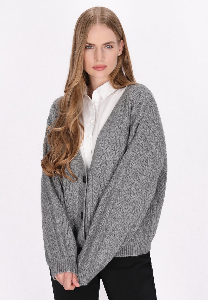 Grijze gebreide cardigan met een gestructureerd herringbone patroon, oversized pasvorm, geribde manchetten en zwarte knopen, gelaagd over een witte blouse.