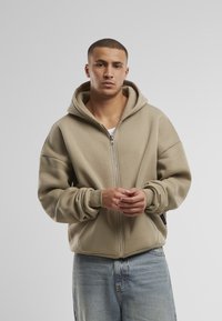 Beige zip-up hoodie met een grote capuchon, ontspannen pasvorm en geribde manchetten, gecombineerd met lichtblauwe beschadigde spijkerbroeken.
