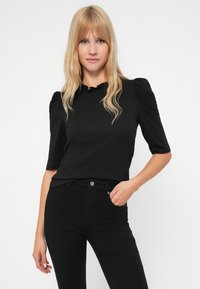 Femme blonde aux cheveux ondulés portant un haut noir à manches bouffantes et un pantalon noir taille haute, posant avec une main dans la poche.