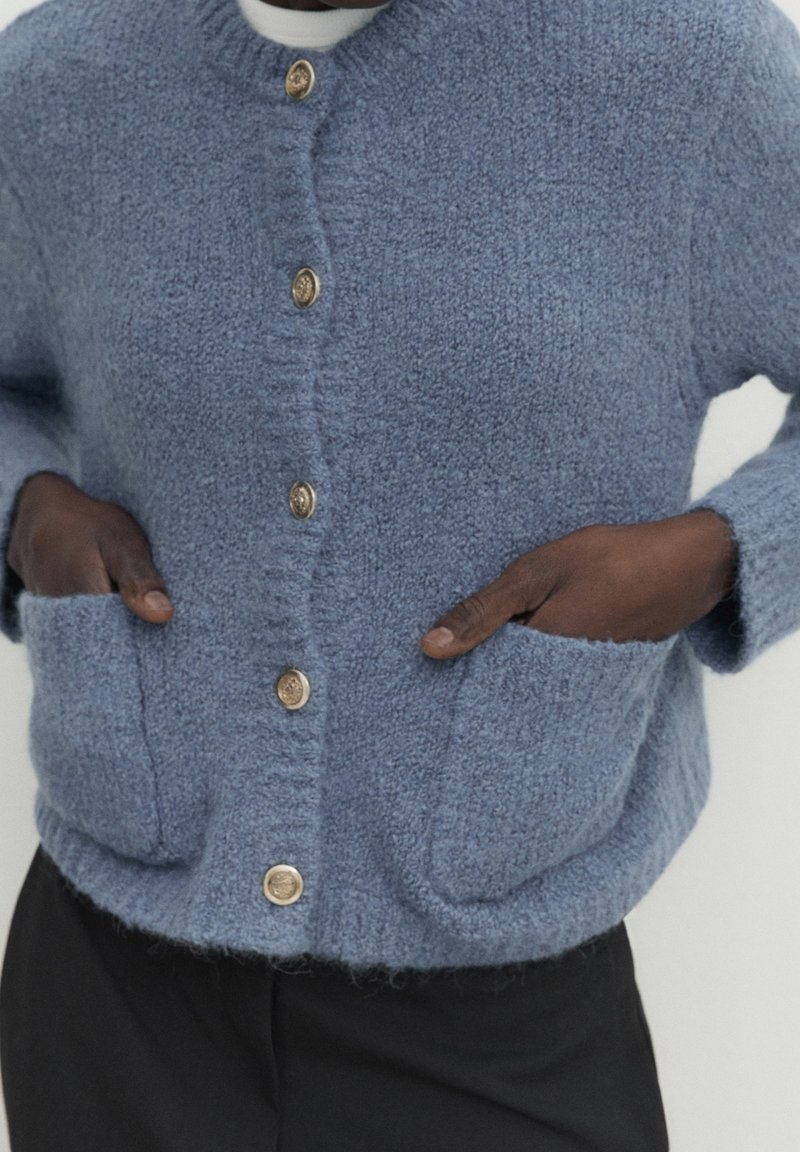Cardigan en tricot bleu avec une finition texturée, doté de cinq boutons dorés, de deux poches avant et d'un col rond.