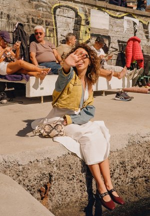 Młoda kobieta w białej sukience i kurtce siedzi na kamiennym murku nad wodą, osłaniając twarz przed słońcem; w tle na ławce siedzą cztery osoby.