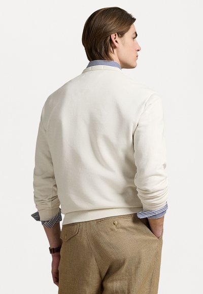 Sweatshirt de culoare crem cu guler rotund și manșete ribbate, purtat peste o cămașă cu dungi albastre și albe, asociat cu pantaloni cu carouri maro.