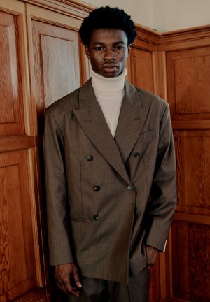 Junger schwarzer Mann, der einen braunen Doppelreiher-Anzug und einen weißen Rollkragenpullover trägt, steht vor holzverkleideten Wänden.