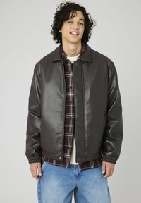 Veste en simili cuir marron avec une fermeture à pression, poignets côtelés et surface texturée, portée sur une chemise à carreaux, accompagnée d'un jeans en denim clair.