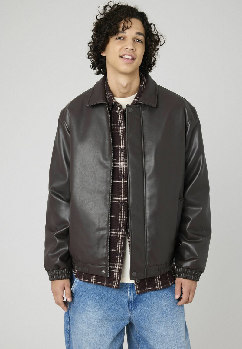 Veste en simili cuir marron avec une fermeture à pression, poignets côtelés et surface texturée, portée sur une chemise à carreaux, accompagnée d'un jeans en denim clair.