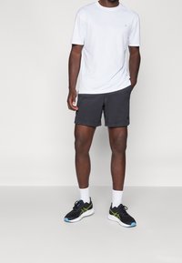 Man står klädd i en vit kortärmad skjorta, mörkgrå shorts, vita strumpor och svarta löparskor med blå detaljer.