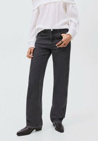 Jeans neri a gamba larga con una finitura testurizzata, abbinati a una blusa bianca off-shoulder. Il modello indossa stivali neri con punta quadrata.