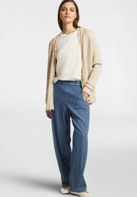 Cardigan beige testurizzato con dettagli in frange sopra una maglietta bianca, abbinato a pantaloni di jeans blu larghi. Calzature sono mocassini di colore chiaro.