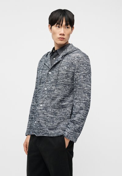 Νέος άνδρας με μαύρα μαλλιά, φοράει ένα μπλε-γκρι μαλλινο πουλόβερ με κουμπιά πάνω από μαύρο πουκάμισο και μαύρο παντελόνι, στέκεται με τα χέρια στις τσέπες.