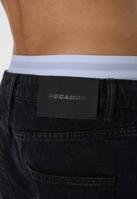 Sorte denimbukser med en grå og lyseblå stribet elastisk talje, der har en sort lædermærke præget med "PEGADOR."