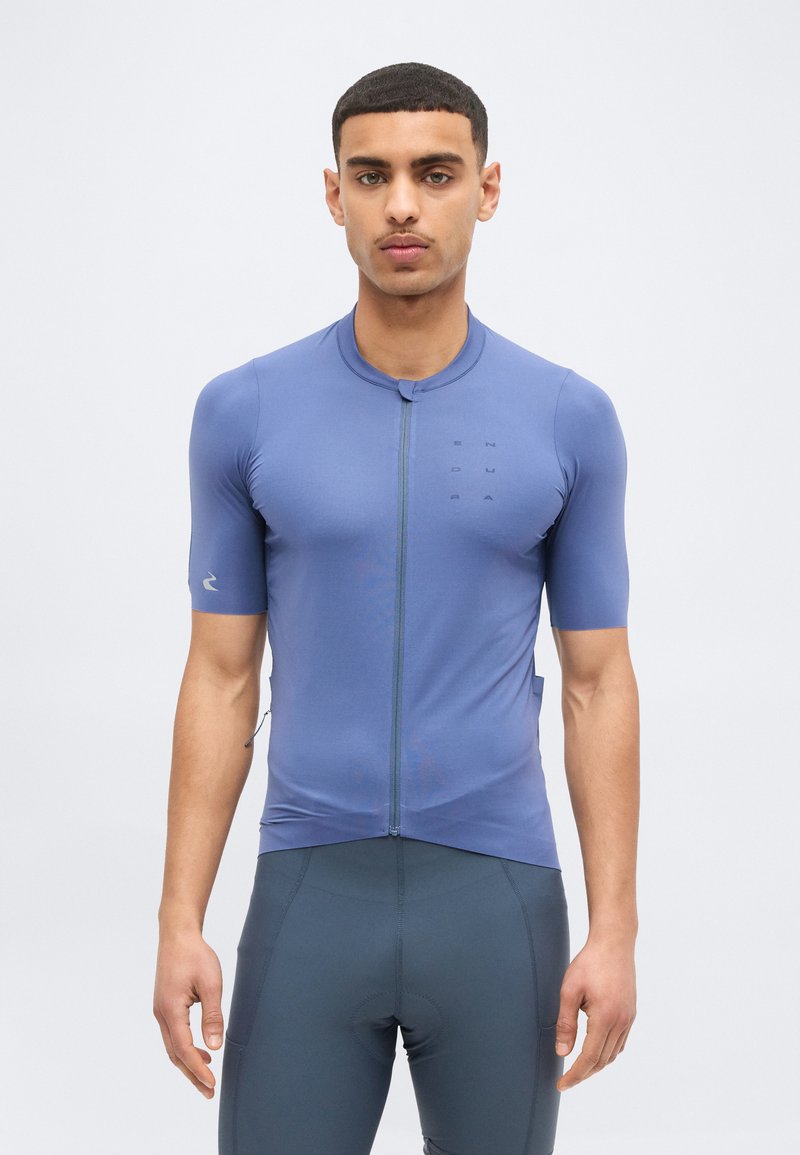 Νέος άνδρας με εφαρμοστό μπλε ποδηλατικό μπλουζάκι με κοντά μανίκια και σκούρο ποδηλατικό σορτς, στέκεται μπροστά σε απλό φωτεινό φόντο.