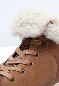 Botte en cuir marron avec une doublure en fourrure crémeuse et duveteuse, présentant des œillets ronds et des lacets plats. La surface texturée ajoute du détail.