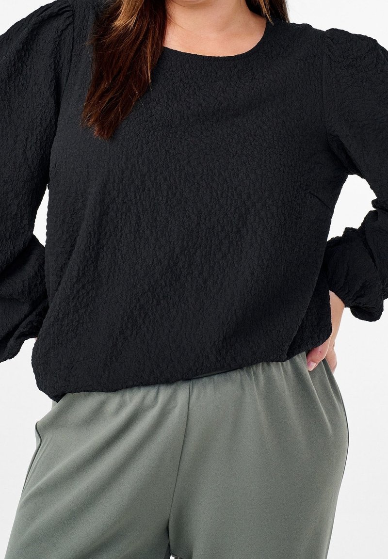 Femme portant un chemisier noir texturé à manches longues et un pantalon ample gris-vert, une main reposant sur la hanche.
