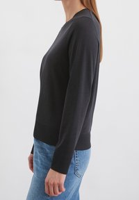 Žena v černém vypasovaném svetru s dlouhým rukávem a modrých džínách stojící bokem proti jednoduchému bílému pozadí.