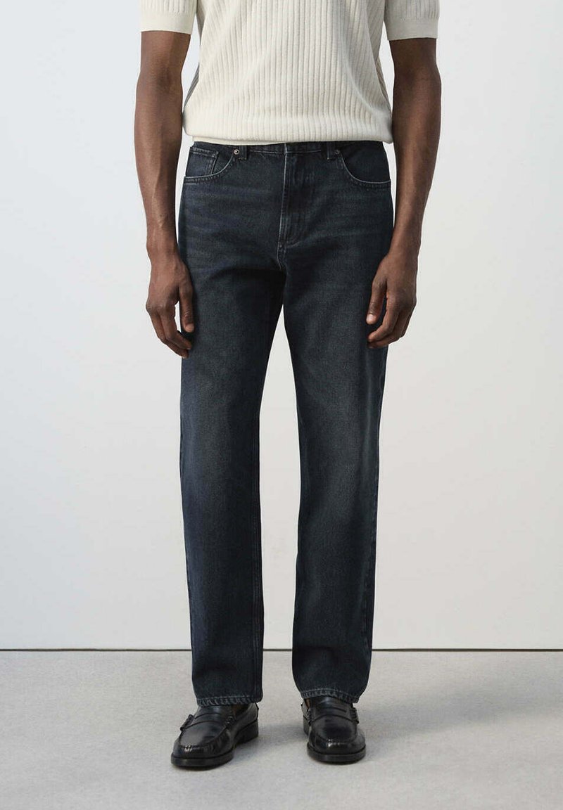 Homme portant un jean bleu foncé droit, un pull blanc côtelé à manches courtes et des mocassins en cuir noirs, debout devant un fond uni.