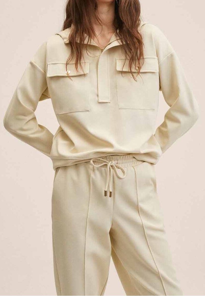 Femme portant une chemise à manches longues de couleur crème avec des poches poitrine boutonnées et un pantalon à cordon assorti, debout les mains sur les hanches.