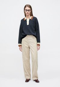 Μπλε ναυτικό πουλόβερ με ριγέ μοτίβο, κρεμ γιακά και καφέ λεπτομέρειες, σε συνδυασμό με ανοιχτό μπεζ παντελόνι κοτλέ με κάθετες ραβδώσεις. Καφέ μοκασίνια.