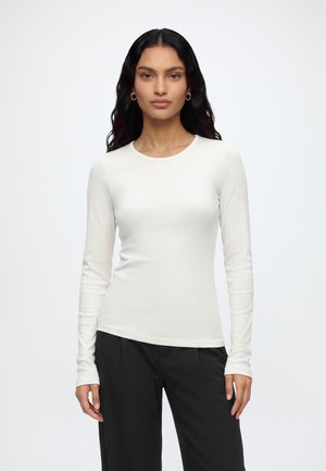 Femme aux longs cheveux foncés portant des boucles d'oreilles créoles dorées, une chemise blanche à manches longues et un pantalon noir, debout devant un fond clair uni.