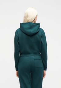 Άτομο με μακριά ξανθά μαλλιά που φοράει σκούρο πράσινο φούτερ με κουκούλα και παντελόνι που ταιριάζει, στέκεται με την πλάτη προς την κάμερα.