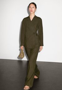 Olijfgroene linnen wikkeljas met een strikceintuur, gecombineerd met bijpassende wijde pantalon. Accessoires omvatten een geweven handtas en platte sandalen.