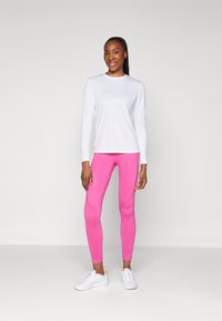 Bela športna majica z dolgimi rokavi v kombinaciji z roza легинсами s teksturiranimi detajli, obuta z belimi supergami. Preprost, prilegajoč se dizajn.