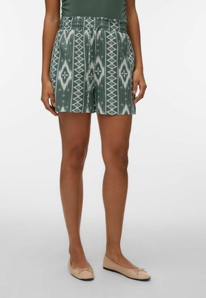 Kvinde iført grønne mønstrede shorts med høj talje og beige ballerinasko, stående mod en ensfarvet hvid baggrund.