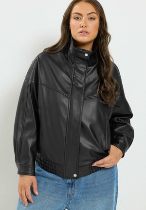 Femme aux longs cheveux bruns portant une veste en cuir noire et un jean bleu, debout devant un fond blanc uni.