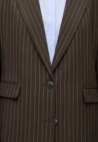 Veste de costume marron à fines rayures avec deux boutons noirs et poches à rabat, portée sur une chemise bleu clair à col.