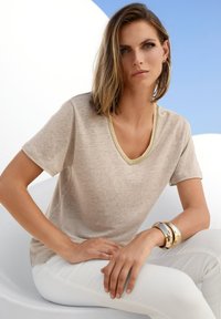 Beige V-Ausschnitt-T-Shirt mit kurzen Ärmeln und einem kontrastierenden Ausschnitt, kombiniert mit weißen Hosen. Die Accessoires umfassen metallische Armreifen.