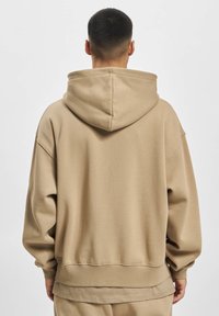 Beige Kapuzenpullover mit lockerem Schnitt, fallenden Schultern und gerippten Bündchen. Glatte Textur und breites Saumdetail.