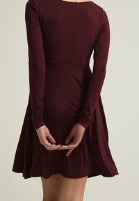Burgundowa sukienka z długim rękawem, dopasowanym bodicem, rozkloszowanym dołem i marszczeniem przy nadgarstkach. Gładki, elastyczny materiał z okrągłym dekoltem.