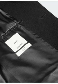 Intérieur d'un blazer en laine noire avec une doublure satinée lisse. Étiquette d'entretien indiquant "MNG", "Coupe slim" et "Fabriqué au Cambodge".