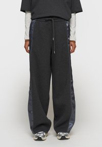 Pantalon de survêtement gris avec des panneaux latéraux en satin, design à jambe large et taille avec cordon de serrage ajustable. Présente une broderie de logo subtile à l'avant.
