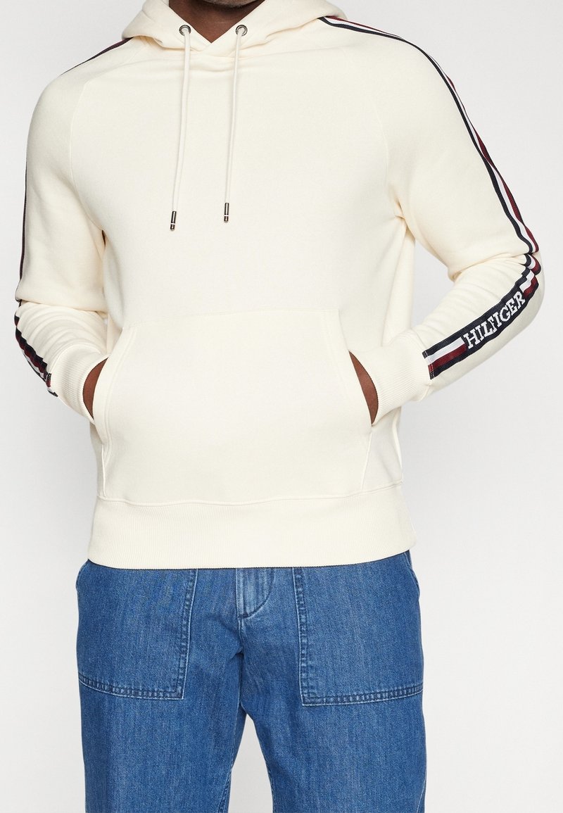 Homme portant un sweat à capuche écru avec des rayures rouges, blanches et bleues et le texte « Hilfiger » sur les manches, associé à un jean bleu avec poche patch.