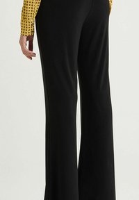 Un pantalon noir évasé présente une texture lisse et une ceinture, associé à un haut jaune à motifs circulaires.