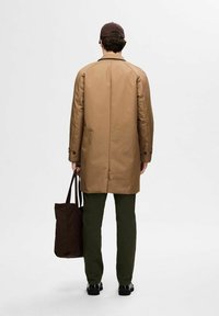 Cappotto marrone chiaro con taglio dritto, abbinato a pantaloni verde oliva. L'individuo tiene una borsa a tracolla marrone scuro. Scarpe nere completano l'outfit.