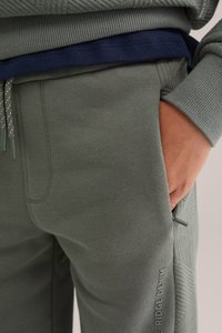 Zelené bavlnené tepláky s hladkou textúrou, bočnými vreckami a rebrovanými akcentmi. Obsahujú detail sťahovacej šnúrky a značkovanie na nohe.