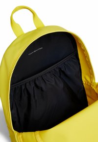 Mochila de tela amarilla con forro de nailon negro, cierre con cremallera y un logotipo en el interior. Presenta un bolsillo elástico para una mejor organización.