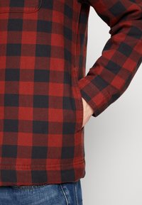 Camisa de flanela em xadrez vermelho e azul-marinho com um bolso frontal, bainha arredondada e detalhes de costura visíveis. O tecido parece suave e ligeiramente texturizado.