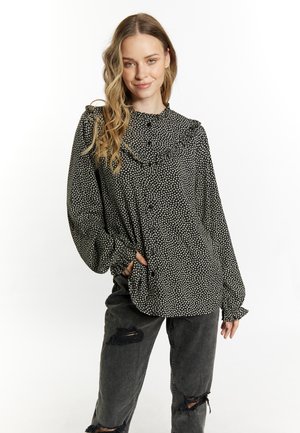Frau mit langen welligen Haaren, die eine schwarze Blumenbluse mit Rüschen und dunkle, zerrissene Jeans trägt, steht mit einer Hand in der Tasche vor weißem Hintergrund.