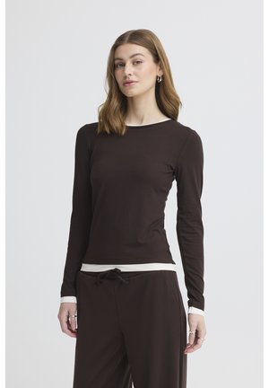 Woman wearing a fitted black long-sleeve shirt and matching black pants with drawstring waist, standing against a plain light gray background.