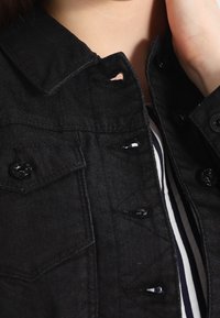 Svart denimjacka med klassisk krage, framknäppning och två bröstfickor. Har ett litet hål vid kragen.