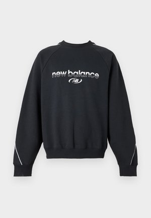 Ung mand med flettet hår iført en sort New Balance sweatshirt og grå New Balance bukser, stående mod en ensfarvet baggrund.