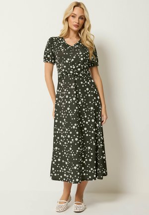 Donna bionda che indossa un vestito midi verde scuro con pois bianchi, maniche corte, scollo a V e scarpe basse bianche intrecciate, in piedi contro un muro semplice.